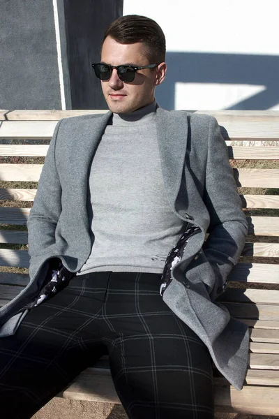 Όμορφος άντρας με φθινοπωρινό παλτό και γυαλιά ηλίου κάθεται σε ένα δρόμο της πόλης. Μενς Μόδα. Ενδύματα και αξεσουάρ. — Φωτογραφία Αρχείου