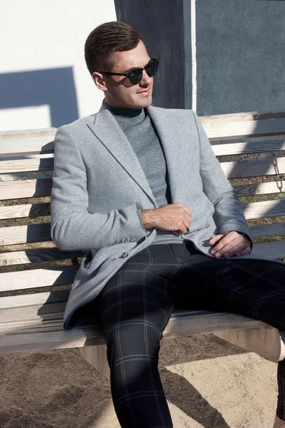 Schöner Mann in Herbstmantel und Sonnenbrille sitzt auf einer Straße in der Stadt. Herrenmode. Bekleidung und Accessoires. Stockbild