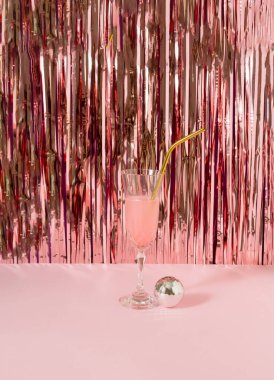 A glass of  pink drink with disco ball against shiny rose gold tassles.