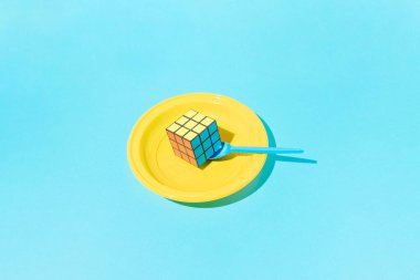 Rubik's cube in yellow plastic plate with blue plastic spoon on blue background.
