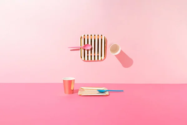 İki altın ve beyaz çizgili kağıt tabak, pembe ve mavi kaşıklar ve pembe arka planda gül sarısı kağıt bardaklar. Asgari konsept.
