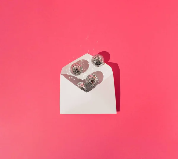 Opened white invitation / letter envelope full of disco balls on pink background. Sharp shadow. Minimal concept.