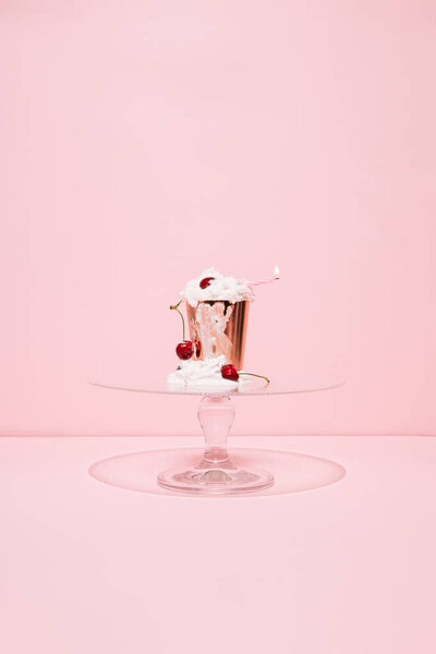 Golden paper with melting whipped cream, cherries and burning candle  on glass cake stand against pink background. Summer cupcake. Minimal concept.