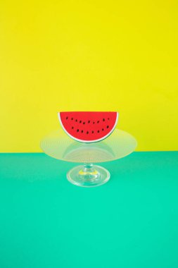 Turkuaz ve sarı arka planda cam pasta standında plastik karpuz dilimi. Asgari yaz konsepti.