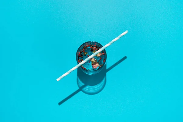 Drink in glass with dried roses, red peppers and striped paper straw on blue background.