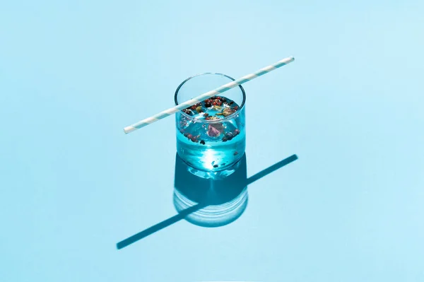 Drink in blue glass with dried roses, red peppers and striped paper straw on blue background.