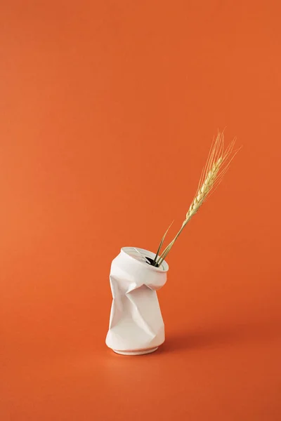 Ear of dried golden wheat in white crumpled beverage can on orange background.