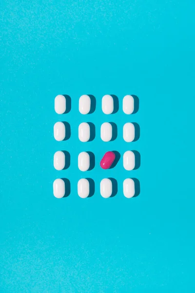 White pills with one tilted pink pill arranged on blue background. Flat lay. Minimal medical concept.