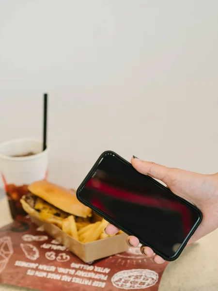 Hand holding blank phone in front of burger set meal with fries and drink