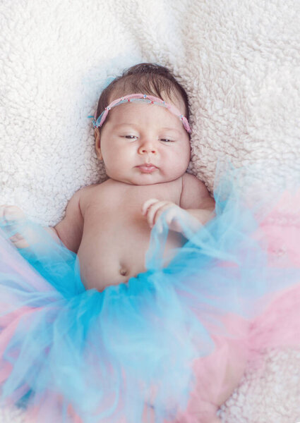  Portrait of a newborn girl with a pink-and-blue skirt and hair decoration rim on the head. Gentle photo with natural light.