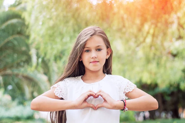 El sevgisi. Sıcak ve güneşli bir yaz gününde parkta kalp sembolü gösteren güzel bir kız. Güneş ışığı etkisi. Mutlu çocukluk kavramı. Aşk sembolü. Ellerle kalp şekillendiren kız