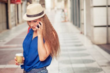 Şapkalı, uzun saçlı, şehir dışında akıllı telefon kullanan ve elinde sıcak kahve içen şehirli bir kızın portresi.. 