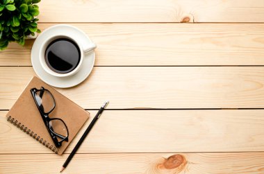 Üst kat ofis masası. Çalışma alanı boş, ofis malzemeleri, kalem, yeşil yaprak ve ahşap zemin üzerinde kahve fincanı.