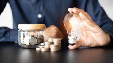 Enerji ve para tasarrufu kavramı. Tasarruf ya da yatırım fikri. Kumbaranın yanında ampul tutan bir iş adamı ve elinde not defteriyle masanın üstünde duran bozuk paralar..