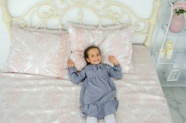 5 yaşında bir kız, güzel, işlenmiş demir bir yatakta, yüzünde neşeli bir ifadeyle yatıyor.