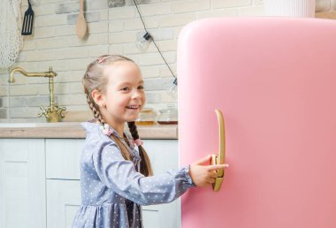 Pembe montlu beş yaşında bir kız eski moda pembe bir buzdolabını eski moda bir mutfakta açıyor..