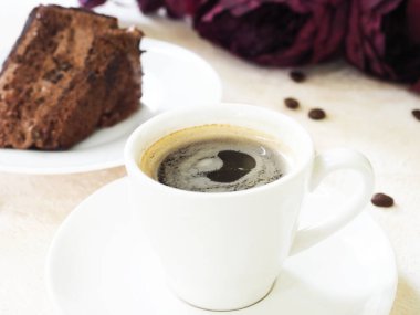 Beyaz bir masada beyaz bir kahve fincanında Espresso kahvesi. Arkasında bir dilim çikolatalı kek var, tatlı olarak..