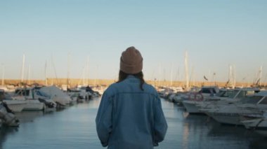 Genç, güzel bir kadın yat kulübünün iskelesinde duruyor, gün batımında rıhtımda, hayal kuruyor, düşünüyor, kameraya mutlu ve nazikçe gülümsüyor, seyahatte ya da şehir keşiflerinde rahat giysiler giyiyor.
