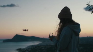 Genç bayanın siluet tutuş görüntüsü, joystick kullanarak helikopteri kontrol etmek, çarpıcı günbatımında drone uçurmak, rüzgarlı doğal arka planda fotoğraf çekmek, seyahat hobisi blogu için videolar, web sitesi