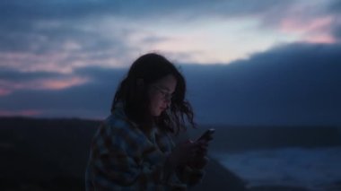Genç rüya gibi milenyum kadınlarının bakış açısı. Cep telefonu kullanan bir kadın, akıllı telefondan sosyal medya haberlerini kaydırırken, yumuşak sinematik ışık yüzüne ve rüzgarlı, renkli pembe, mavi günbatımı arka planda