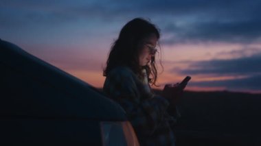 Sinema yumuşak manzarası, bulanık odaklı, akıllı telefon kullanan, gülümseyen, sosyal medyayı kaydıran, hafta sonları kamp arabasına yaslanan, gezen, renkli Porto Günbatımında tatil yapan genç, bin yıllık genç bir kadın.