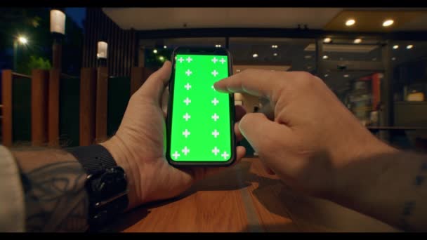 年轻男性纹身手的近照 手表垂直握着 使用绿色屏幕的手机 桌子上滚动的智能手机触摸屏 背景是柔和的咖啡馆灯光 — 图库视频影像