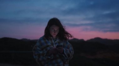 Genç hayalperest kadın cep telefonu kullanıyor, sohbet ediyor, mesajlaşıyor, akıllı telefondan sosyal medya haberlerini süzüyor. Yumuşak sinematik yüz ve rüzgarlı, pembe, arka planda mavi gün batımı.