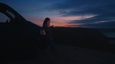 Yumuşak bulanık odak noktası güzel genç kadın silueti akıllı telefon, mesaj, kaydırma sosyal medya kullanarak, sohbet açık havada kamp arabasına yaslanarak karanlık Porto günbatımı renklerinde sıcak farlarla