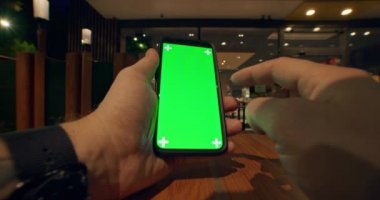 Dövmeli genç erkek elleri, saatleri dikey tutan öğrenci ya da işçiyi ve arka planda koyu yumuşak ışık olan bir kafede otururken yeşil renkli ekranlı cep telefonunu kullanan genç erkek ve işçi görüntülerini kapat.