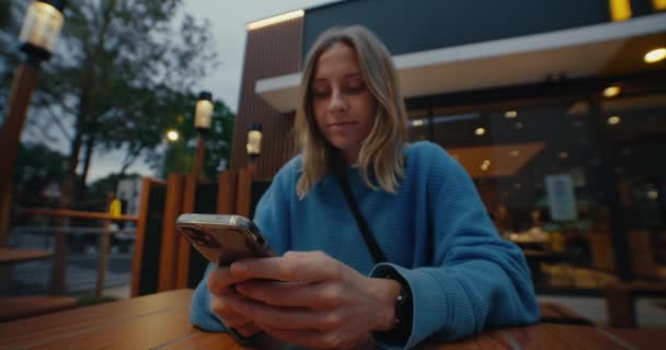 迷人的年轻女子 浅色的头发 身穿蓝色毛衣 坐在咖啡馆外面 用智能手机 发短信 在社交媒体上与朋友聊天 在手机上滚动新闻 — 图库视频影像