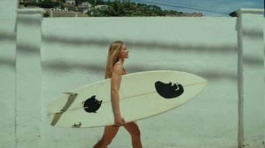 Side tracking shot of young blonde woman walk towards beach with surfboard. Surfing town summertime vacation. Female surfer beach lifestyle. Woman surfer enjoy summer breeze in portugal or california