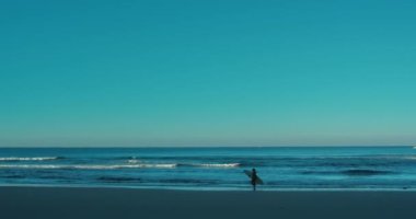 Silhouette of surfer woman walk slowly on edge of water with surfboard. Woman in cold water swimsuit at early morning sunrise surfing session