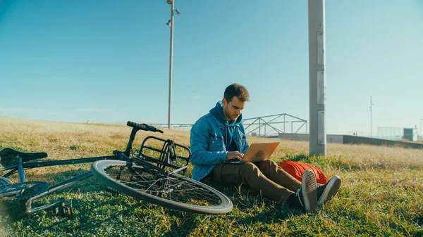 Handsome young man with commuter bicycle sit outside in park, work on laptop. Freelancer or student use free time or lunch break to work on project outdoors. Millennial work habits