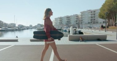 Blond hair young woman walks barefoot with skateboard in her hand in city centre with boats in the sunny morning. Teenager looks at camera and smiles. Handheld footage with natural lens flare