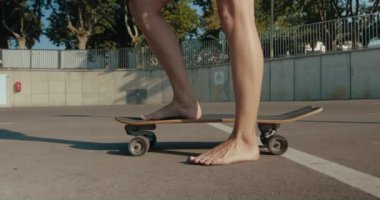 Handheld close-up side view of barefoot woman feet on skateboard slowly moving it front and back ready to skate, summer vacation vibe at golden hour