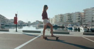 Healthy beautiful woman in white shorts and red shirt moving surfskate front and back with one foot looking at the camera. Blond hair and barefoot young woman poses with skateboard