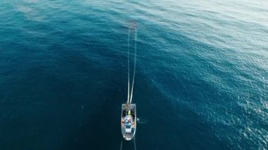 White small fisherman trawler with crew on board. People work together to pull out net with caught sardines from deep blue waters of open sea. Aerial shot with front view
