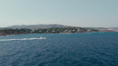 Dolly zoom in aerial shot of coastal town hidden in trees, big luxurious yacht moves fast forward in open sea and leaves splashes and wave foam trace in blue water. Summer chill and vacation in Europe