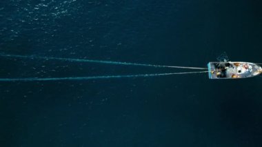 Tracking shot of fisherman team on small trawler, pulling out huge net from deep blue ocean. Top view aerial with object in middle