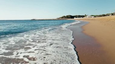 Dolly shot on sea shore with sand beach close to waves with foam at high tide in the early morning next to resort or camping. Summer vacation vibe. Extreme close up