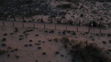 Girl finishes her run workout and looks on smart watch. Fine sand beach with dunes at twilight near shoreline. High quality 4k footage. Orbit drone shot 