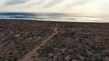 Girl runs fast towards camera between sand dunes at sunset. Cloudy weather on scenic beach next to ocean shoreline. High quality 4k footage Dolly zoom out shot