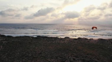 Hava yörüngesi, günbatımında kum tepelerinin üzerinde uçan kırmızı kanatlı paraglider etrafında çekildi. Golden Hour 'da antrenman. Deniz manzaralı ve dalgalı dramatik bulutlu gökyüzü.