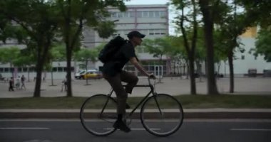 Shady City caddesinde bisikletli bir gencin yan görüntüsü. Modern çağın hippi kıyafetli adamı bisikletinin üstünde pedal çeviriyor. Yeşil ve ekolojik şehir turu 