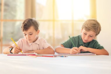 Evde ödev yapan iki çocuk, pastel boyalarla resim çiziyorlar.