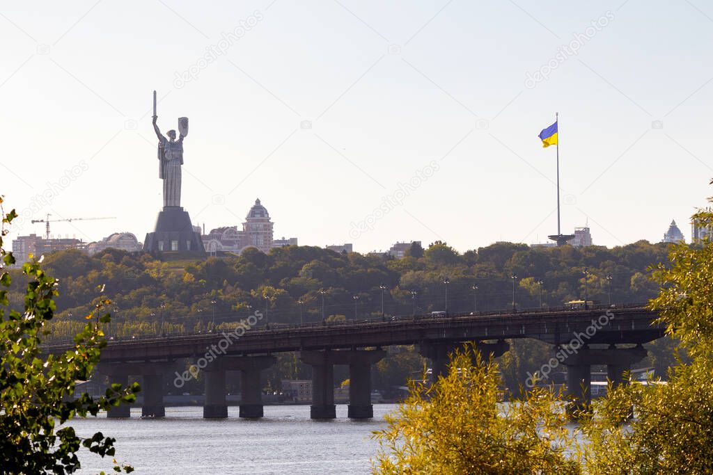 Bright evening cityscape of the big city on the hill over wide river Dnipro in awesome bright sunset in Kyiv, Ukraine. View at largest ukrainian national flag on flagpole and monument Motherland.