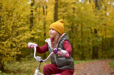 Gülümseyen bir kız sonbahar parkında bisiklet sürüyor.