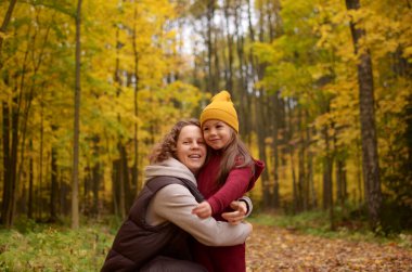 Kıvırcık saçlı gülümseyen bir anne ve sonbahar parkında yürüyen küçük bir kız çocuğu.