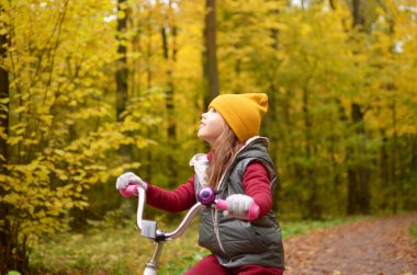 Gülümseyen bir kız sonbahar parkında bisiklet sürüyor.