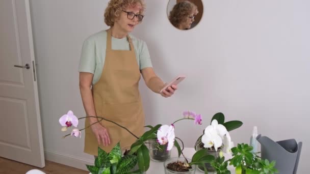 Retrato de mujer mayor en delantal amarillo toma fotos de orquídeas — Vídeo de stock